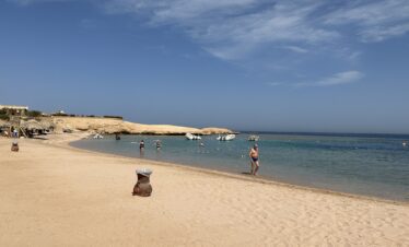 Plage de Sharm el-Naga avec sable, mer, parasols en feuilles et des touristes dans l'eau.