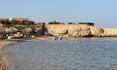 Plage de Sharm el-Naga avec sable, mer, parasols en feuilles et des touristes dans l'eau.