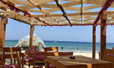 Un restaurant de plage avec des tables et chaises en bois installées directement sur le sable, offrant une vue imprenable sur la mer et une sculpture géante en forme de tortue métallique.