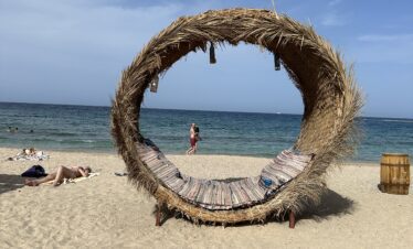 Lit circulaire en bois et paille sur une plage de sable avec vue sur la mer.