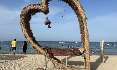 Structure en forme de cœur en bois et paille sur la plage, avec la mer en arrière-plan.