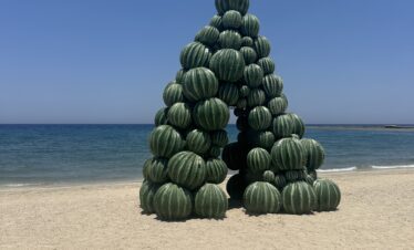 Une structure artistique en forme d’arche composée de pastèques empilées sur une plage de sable, avec la mer en arrière-plan.