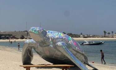 Une sculpture métallique brillante en forme de tortue sur une plage de sable, avec une table de pique-nique au premier plan et la mer en arrière-plan.