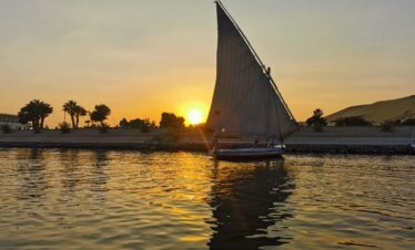 Une felouque navigue paisiblement sur le Nil au coucher du soleil, avec ses voiles déployées et des palmiers en arrière-plan.