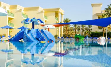 Piscine avec jeux pour enfants, grande grenouille et transats sous les palmiers.