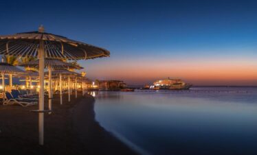 Plage de nuit avec transats, parasols et palmiers, et l’hôtel en arrière-plan.