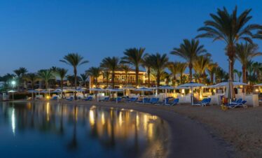 Plage de nuit avec transats, parasols et palmiers, et l’hôtel en arrière-plan.