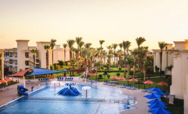 Espace aquatique pour les tout-petits avec petits toboggans, transats, parasols et palmiers, vue sur l'hôtel et un aperçu de la plage au loin.