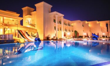 Vue nocturne de la piscine avec espaces pour les tout-petits et les enfants, toboggans, transats et parasols fermés, éclairages illuminant l'hôtel.