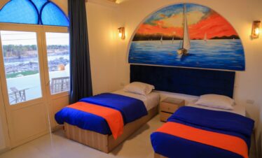 Chambre avec deux lits simples en bois, tête de lit sculptée, grande peinture représentant le Nil au coucher du soleil et vue sur le Nil.