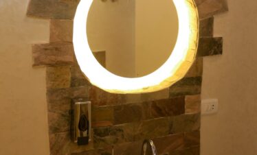 Lavabo blanc avec parement de pierre formant des rayons autour d’un miroir rond lumineux.