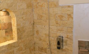 Douche avec parements de pierre dorés, niches murales éclairées et pommeau de douche, avec un aperçu de la fenêtre.