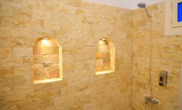 Douche avec parements de pierre dorés, niches murales éclairées et pommeau de douche, avec un aperçu de la fenêtre.