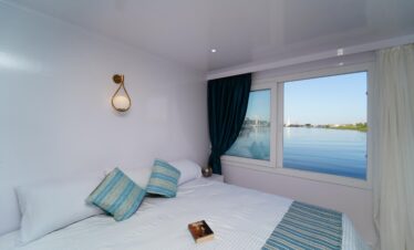 Chambre double minimaliste avec grand lit, tables de chevet et fenêtre latérale donnant sur le Nil.