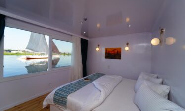 Chambre double minimaliste avec grand lit, tables de chevet et grande fenêtre ouverte sur le Nil.