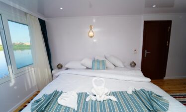 Chambre élégante avec un lit double à bord d’un bateau Dahabiya en Égypte, avec vue sur le Nil.
