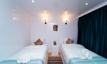 Chambre cosy à bord d’un bateau de croisière sur le Nil, avec deux lits simples, rideaux fermés et une ambiance intime et apaisante.